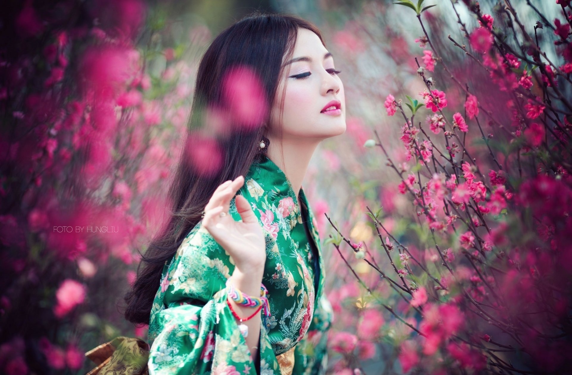HD desktop wallpaper featuring an Asian woman in a green floral kimono, amidst blooming pink flowers. She stands gracefully, eyes closed, exuding serenity and elegance.