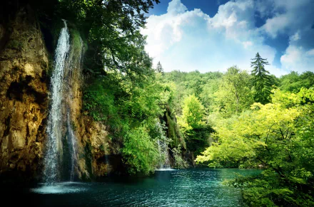 A serene waterfall cascades into a vibrant blue pool, surrounded by lush green trees under a bright sky, creating a stunning nature-inspired HD desktop wallpaper.