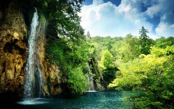 A serene waterfall cascades into a vibrant blue pool, surrounded by lush green trees under a bright sky, creating a stunning nature-inspired HD desktop wallpaper.