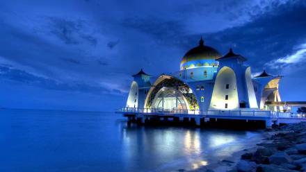 Night view of the illuminated Malacca Straits Mosque in Malaysia, reflecting on calm waters under a deep blue sky, captured as an HD desktop wallpaper background.