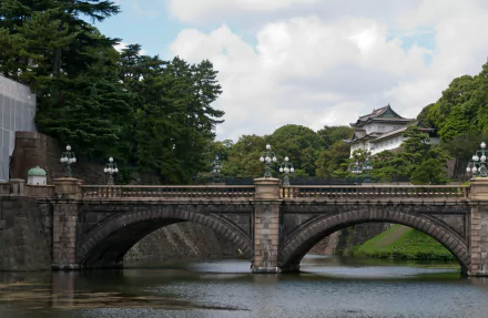 View of a man-made stone bridge over water with lush greenery and a traditional structure in the background at Tokyo Imperial Palace, Japan.