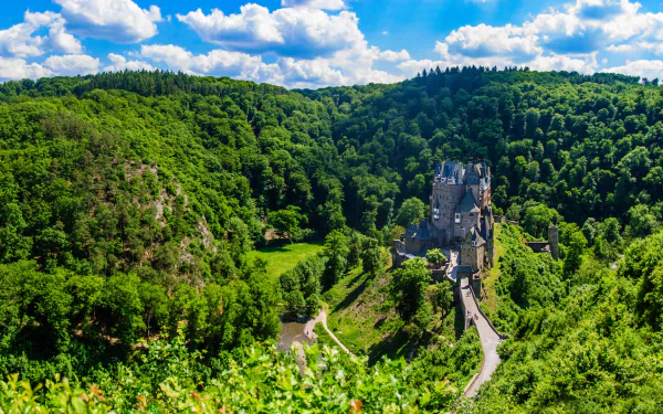 A stunning HD desktop wallpaper of Germany’s Eltz Castle nestled within a dense green forest under a bright blue sky with scattered clouds.