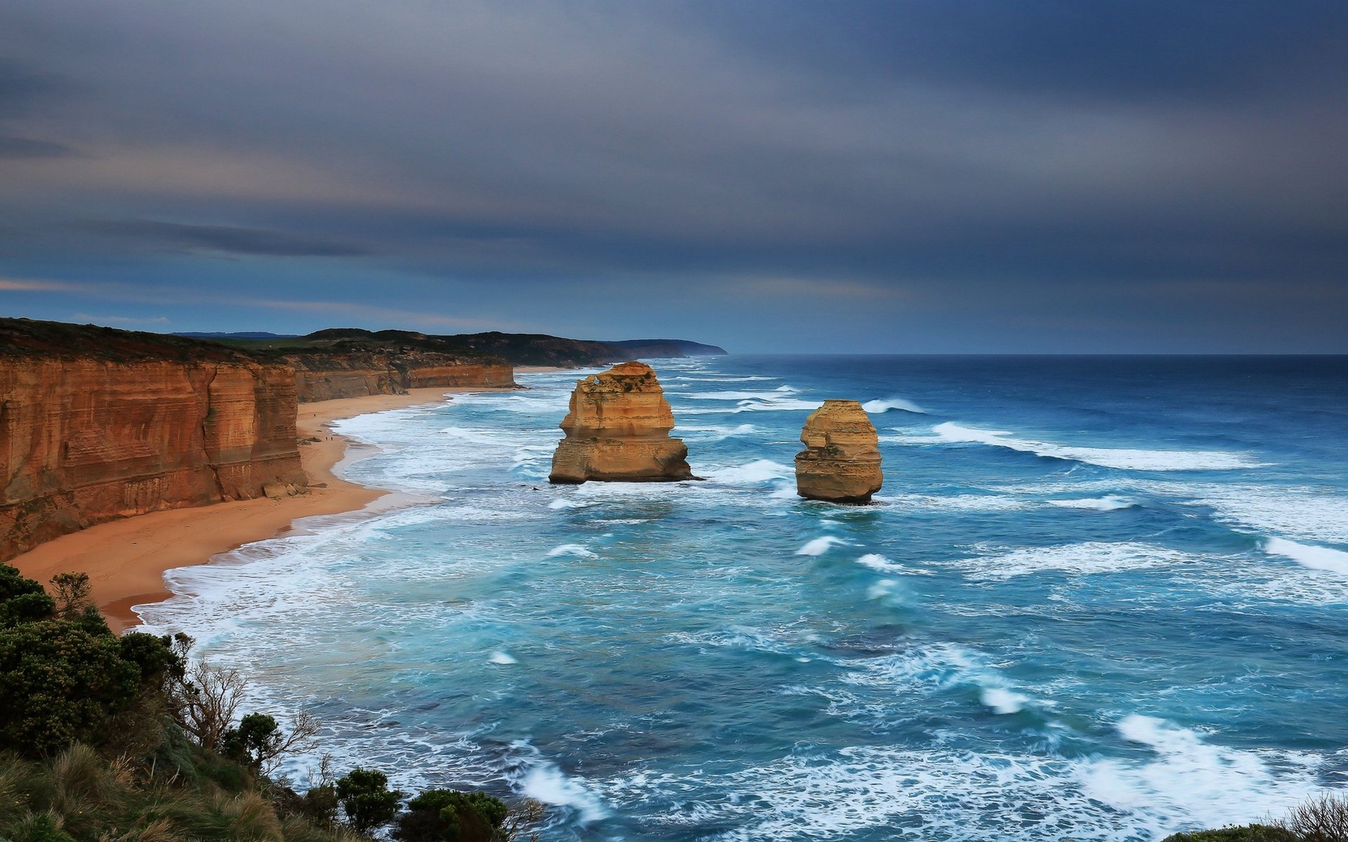 HD PC desktop wallpaper and background: a nature coastline with turquoise surf, two towering sea stacks, golden cliffs and a moody cloudy sky.