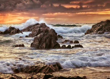 HD PC desktop wallpaper of a rocky shoreline at sunset, waves crashing beneath a cloud-filled sky as the tide sweeps around jagged sea rocks, a dramatic sea and coastal nature scene.