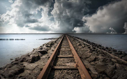 HD wallpaper of railroad tracks extending into the horizon under a dramatic cloud-covered sky, set against a vast body of water.