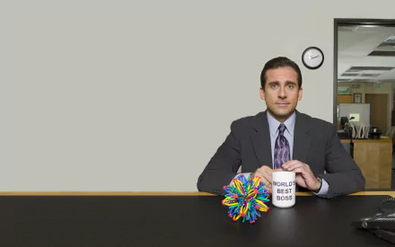 Steve Carell sitting at a desk in a suit, holding a World's Best Boss mug, with a colorful stress ball, from the TV show The Office (US). A minimalistic office background completes the scene.