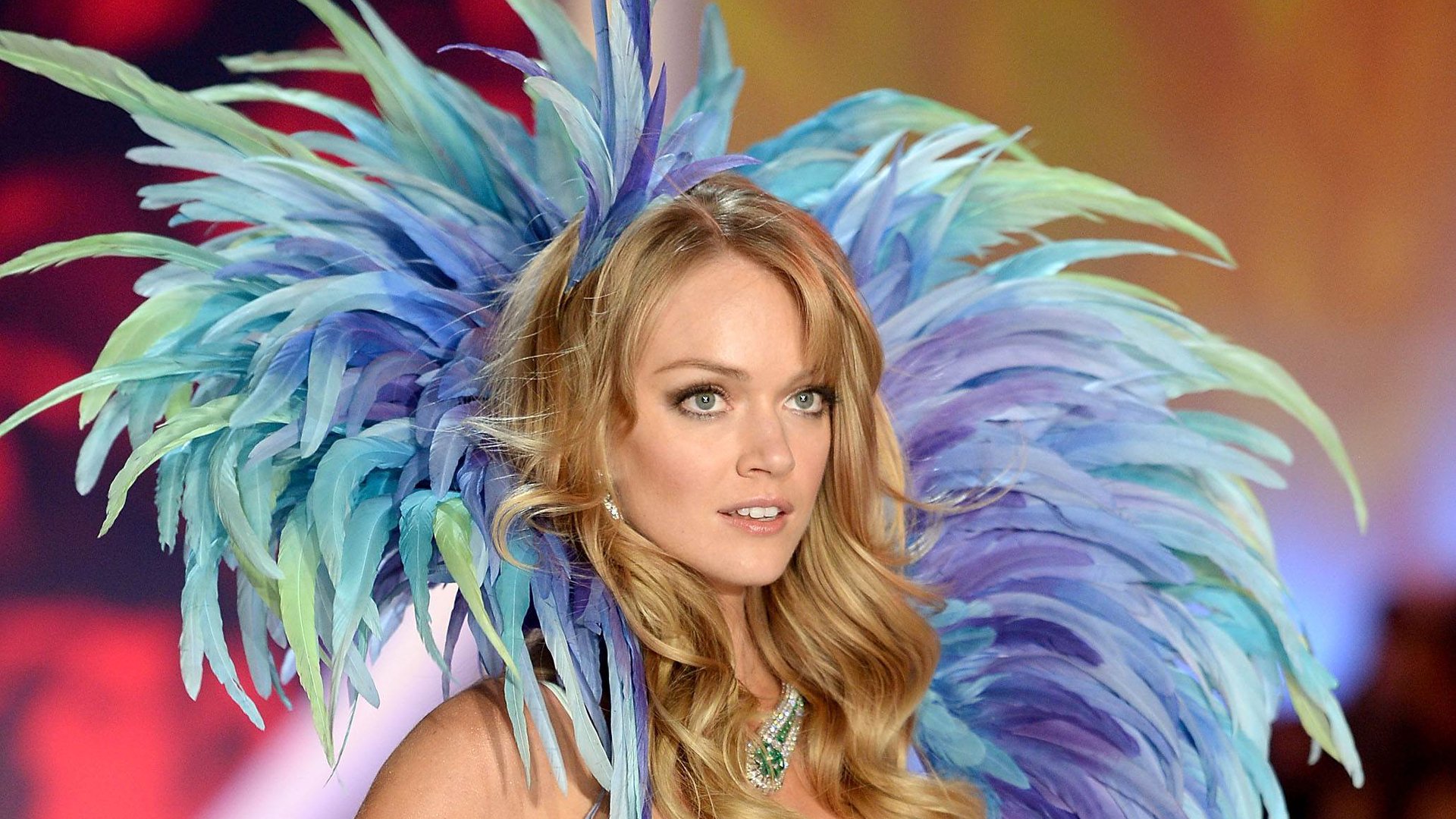 HD desktop wallpaper featuring American model Lindsay Ellingson wearing an elaborate blue and purple feathered headdress against a warm blurred background.
