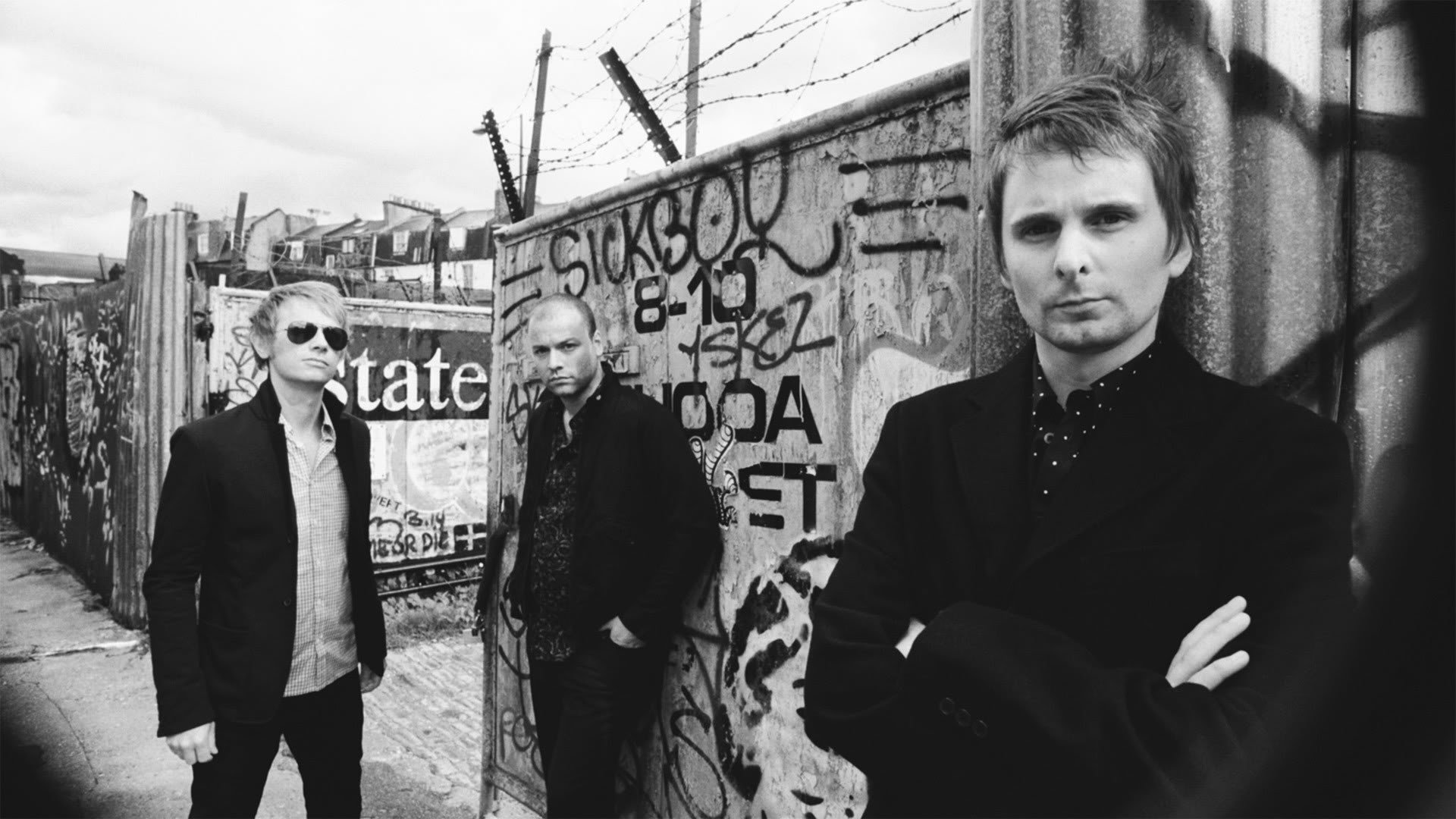 Monochrome HD PC wallpaper of a three-member rock band in suits against a graffiti-covered urban wall; moody music portrait — tags: music, Muse