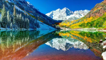 Snow-covered Maroon Bells mountains reflect clearly in the calm waters of Maroon Lake, surrounded by colorful autumn trees in Colorado.