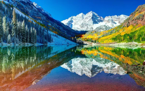 Snow-covered Maroon Bells mountains reflect clearly in the calm waters of Maroon Lake, surrounded by colorful autumn trees in Colorado.