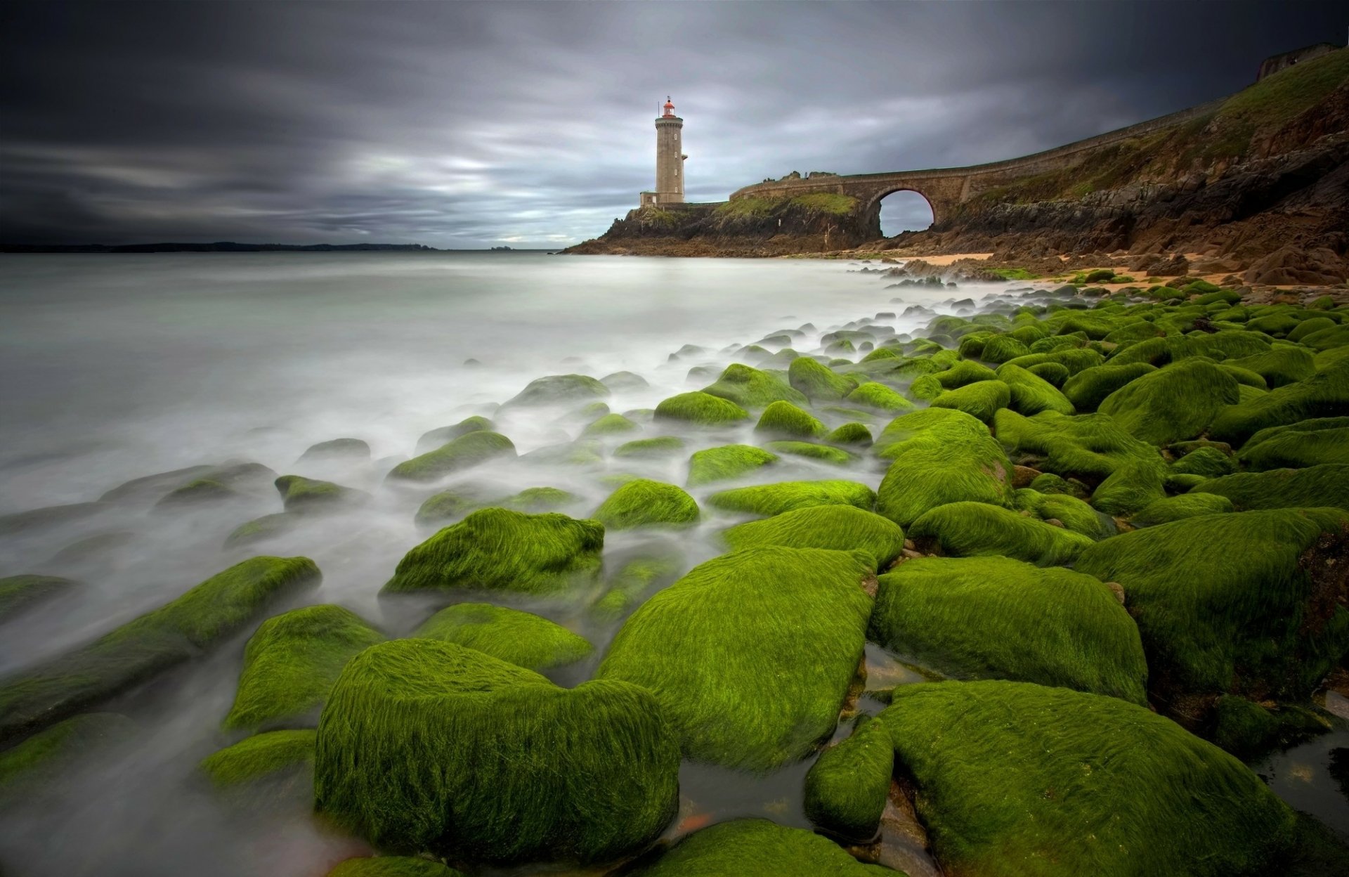 HD desktop wallpaper of a rocky seashore with vibrant green moss, a cloudy sky, and a man-made lighthouse perched on a distant cliff by the water.