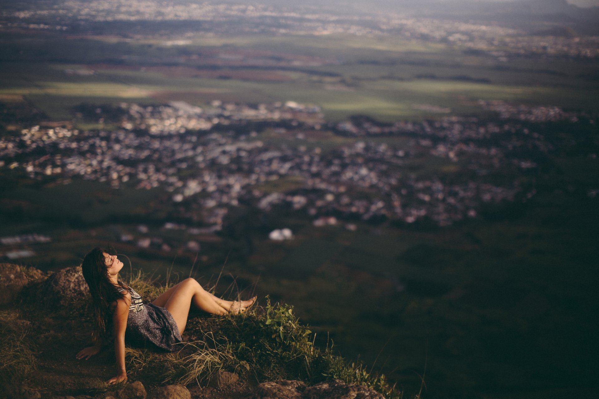 HD PC desktop wallpaper showing a woman sitting on a grassy hill, gazing thoughtfully over a blurred cityscape, capturing a serene, introspective mood.