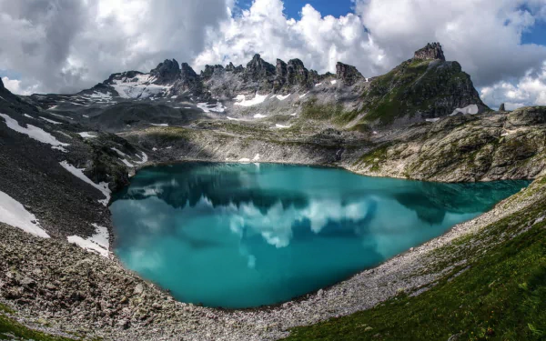A stunning HD desktop wallpaper showcasing a serene alpine lake surrounded by rugged mountain ridges under dramatic cloud-filled skies in the Alps.