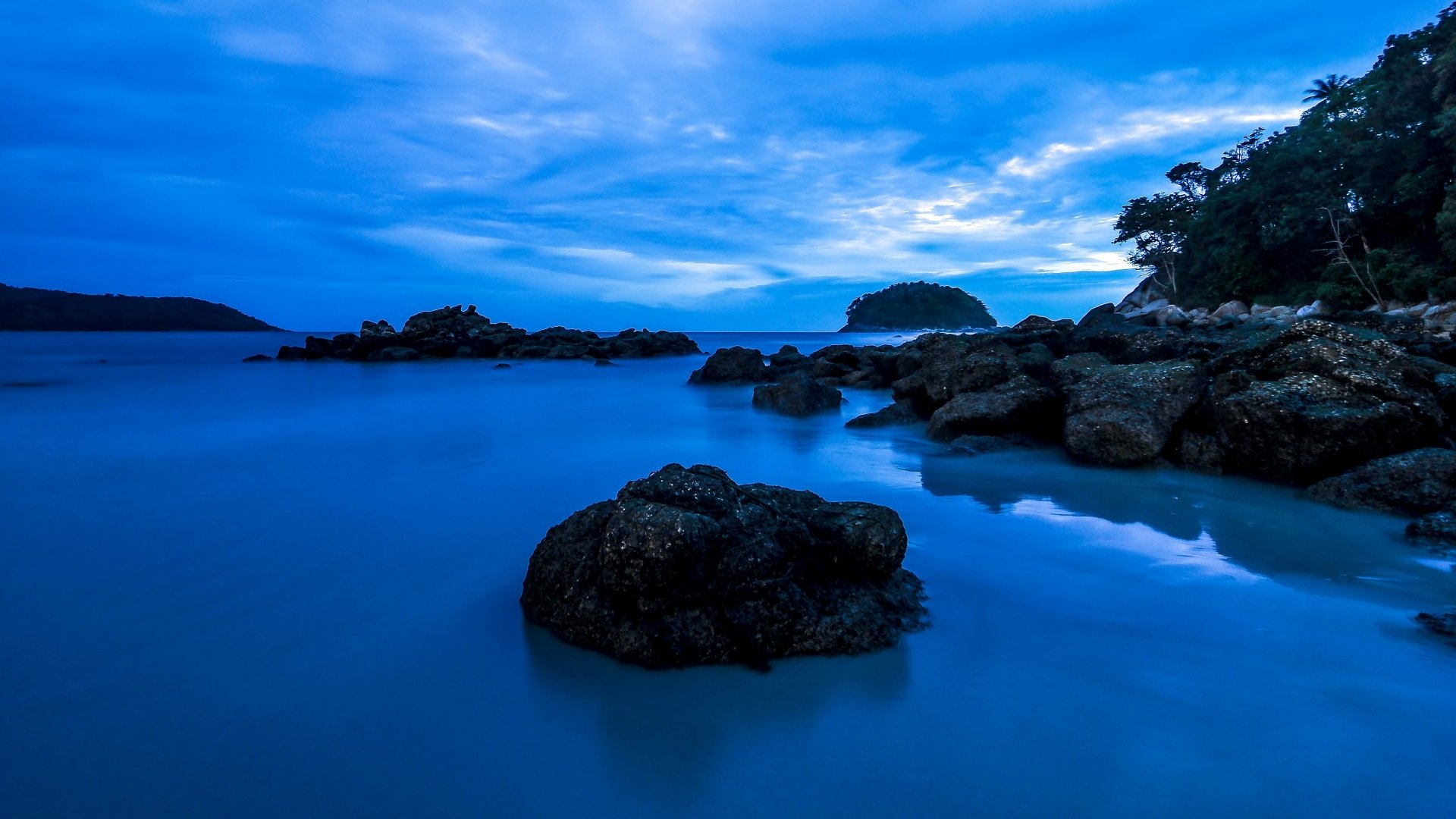 4K Ultra HD desktop wallpaper showing a serene ocean scene with rocks, lush greenery, and a vivid blue sky filled with clouds.