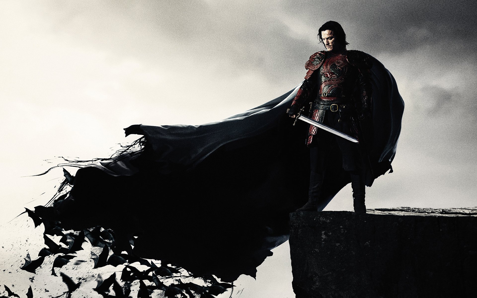 HD desktop wallpaper featuring the movie Dracula Untold. The image portrays Dracula, cloaked and holding a sword, standing on a cliff with bats flying in the background.