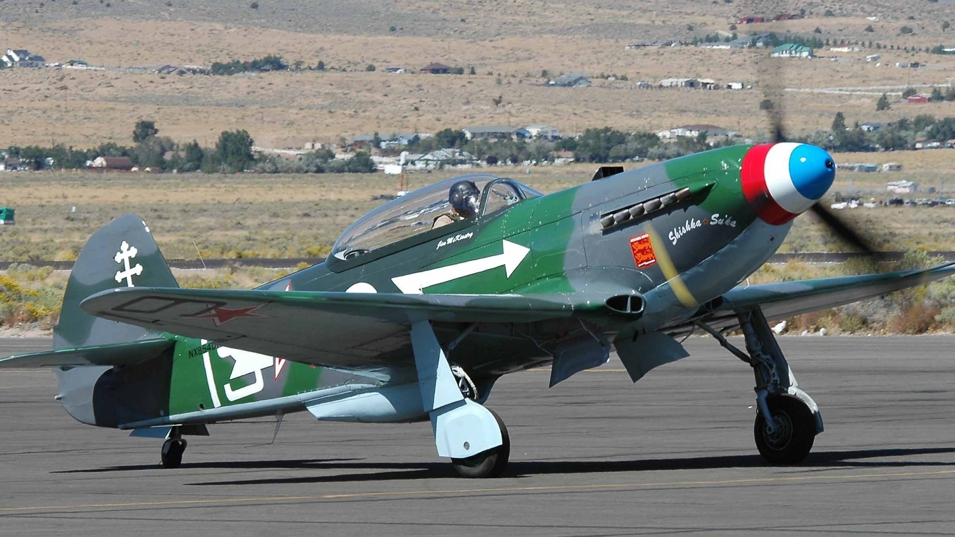 HD PC desktop wallpaper: green military Yakovlev Yak-3 fighter with tricolor nose and white arrow marking taxiing on a runway against an arid landscape background.