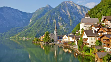 A stunning HD wallpaper of Hallstatt, showcasing charming houses, a church steeple, and lush mountains reflecting in the serene lake. A perfect blend of nature and man-made beauty.