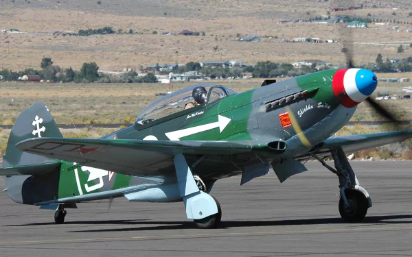 HD PC desktop wallpaper: green military Yakovlev Yak-3 fighter with tricolor nose and white arrow marking taxiing on a runway against an arid landscape background.