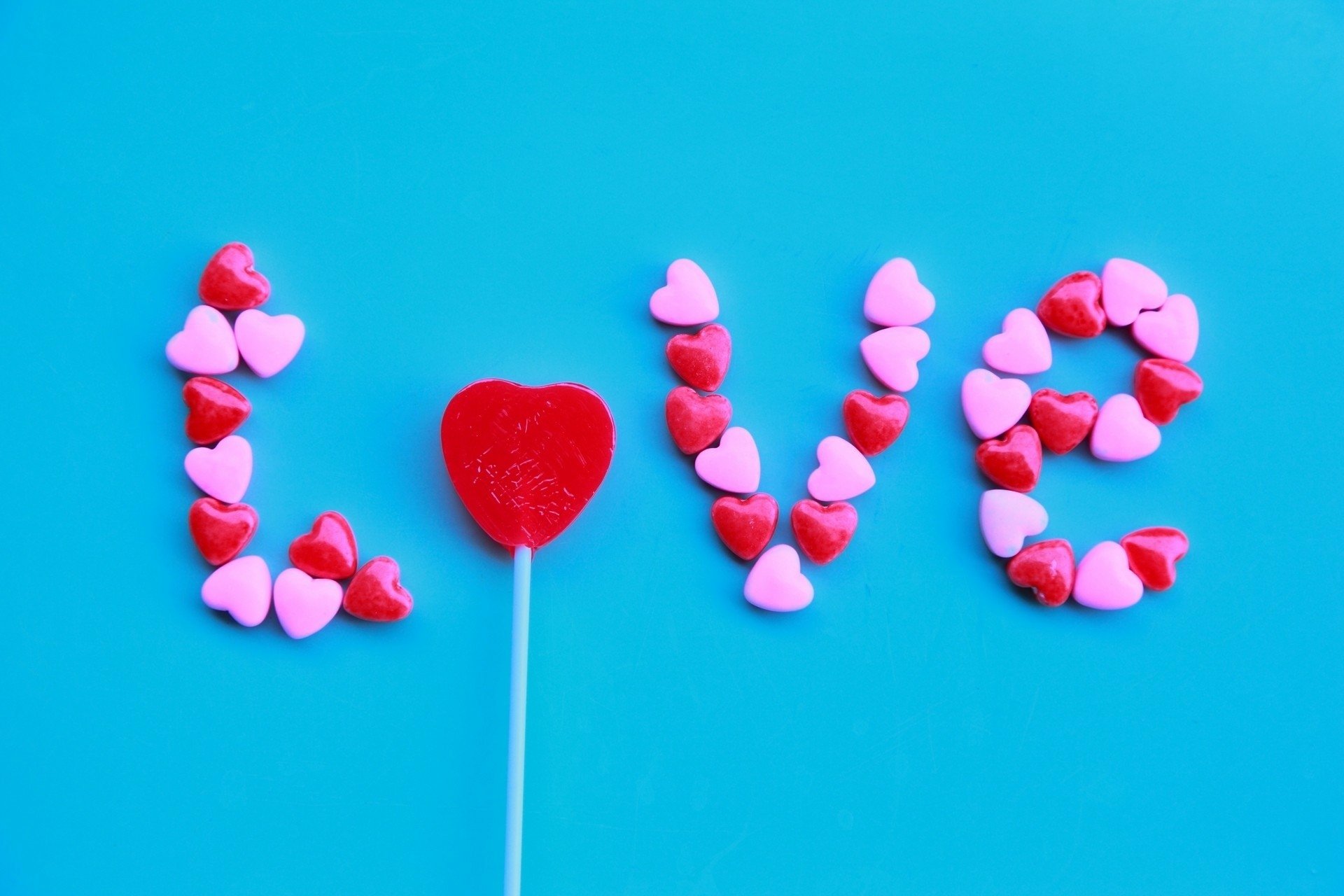 Colorful heart-shaped candies spell out love beside a red lollipop on a vibrant blue background, creating an artistic celebration of affection and sweetness.