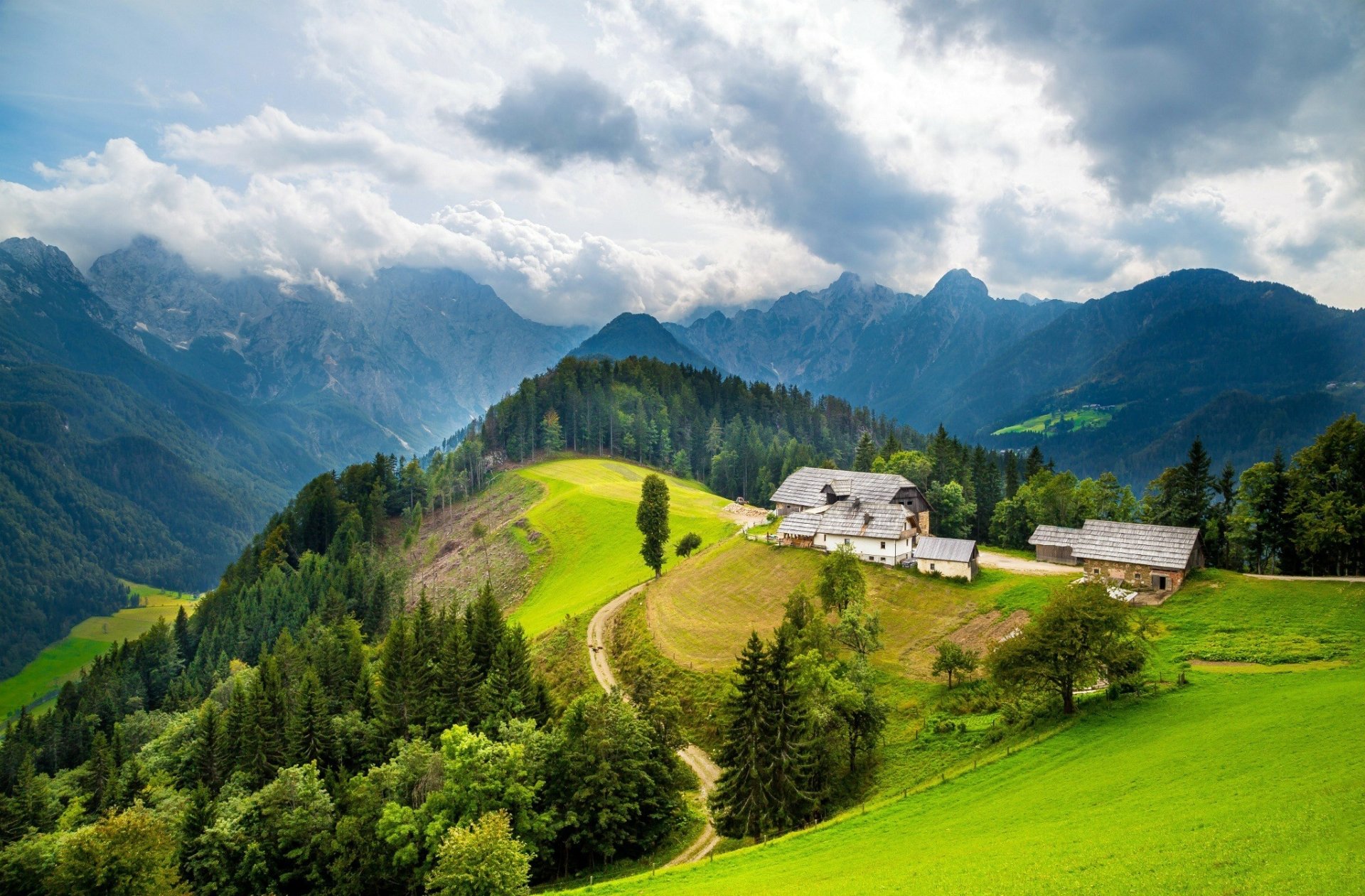 A scenic landscape featuring a winding road leading to a charming house on a hillside, surrounded by lush woodlands and towering mountains under a cloudy sky.