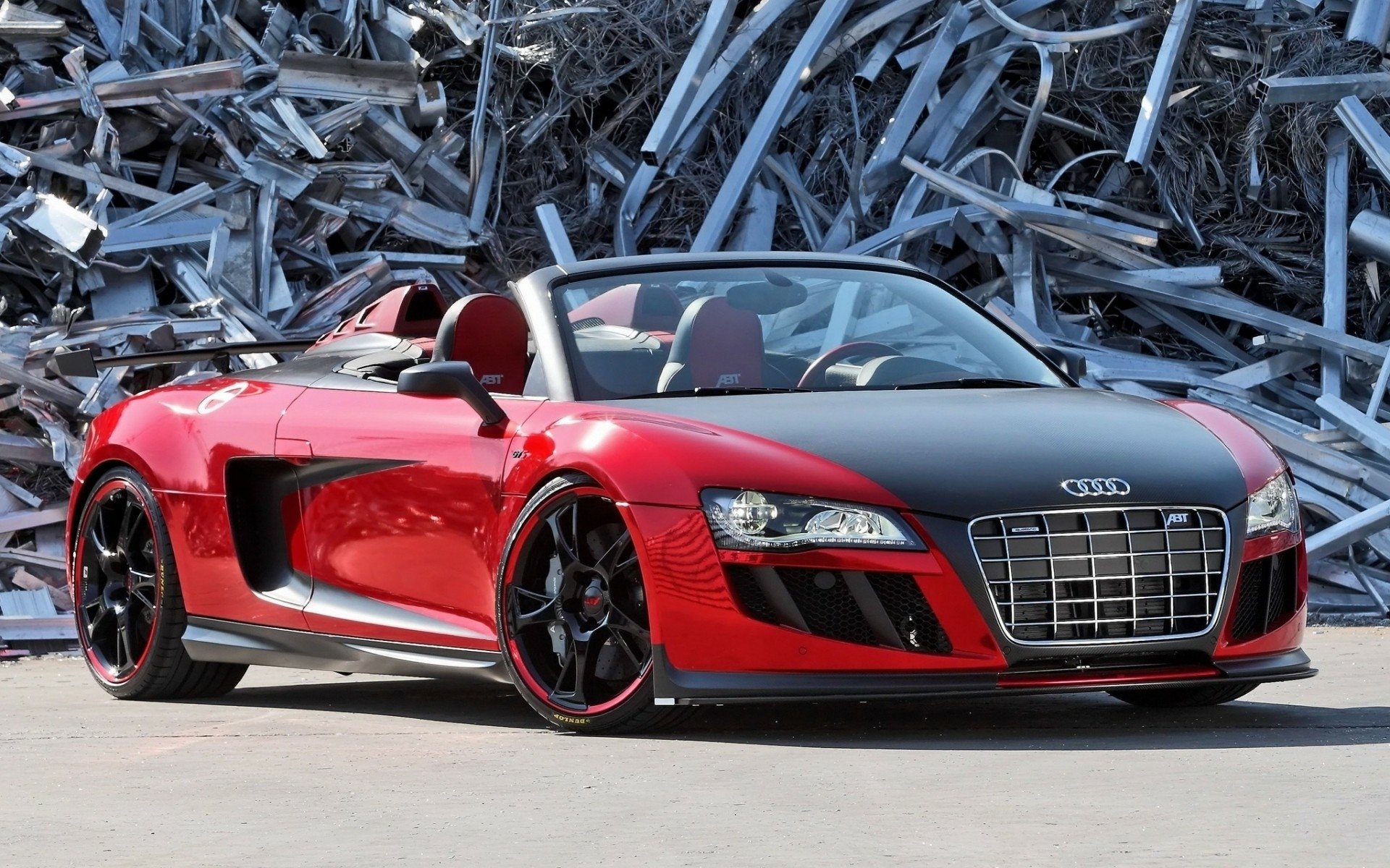HD PC desktop wallpaper featuring a sleek red Audi R8 convertible parked in front of a pile of metal scraps, showcasing the vehicle's sporty design and bold presence.
