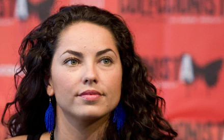 A HD desktop wallpaper featuring Mexican actress Barbara Mori, showcasing her with curly hair and blue earrings against a vibrant red background.