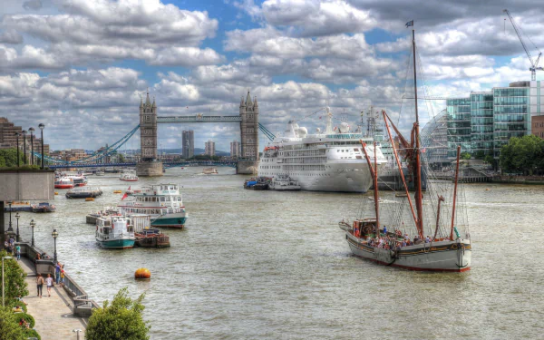HD desktop wallpaper of the River Thames in London with the man-made Tower Bridge, cruise and sailing boats on the river and riverside buildings beneath a dramatic cloudy sky.