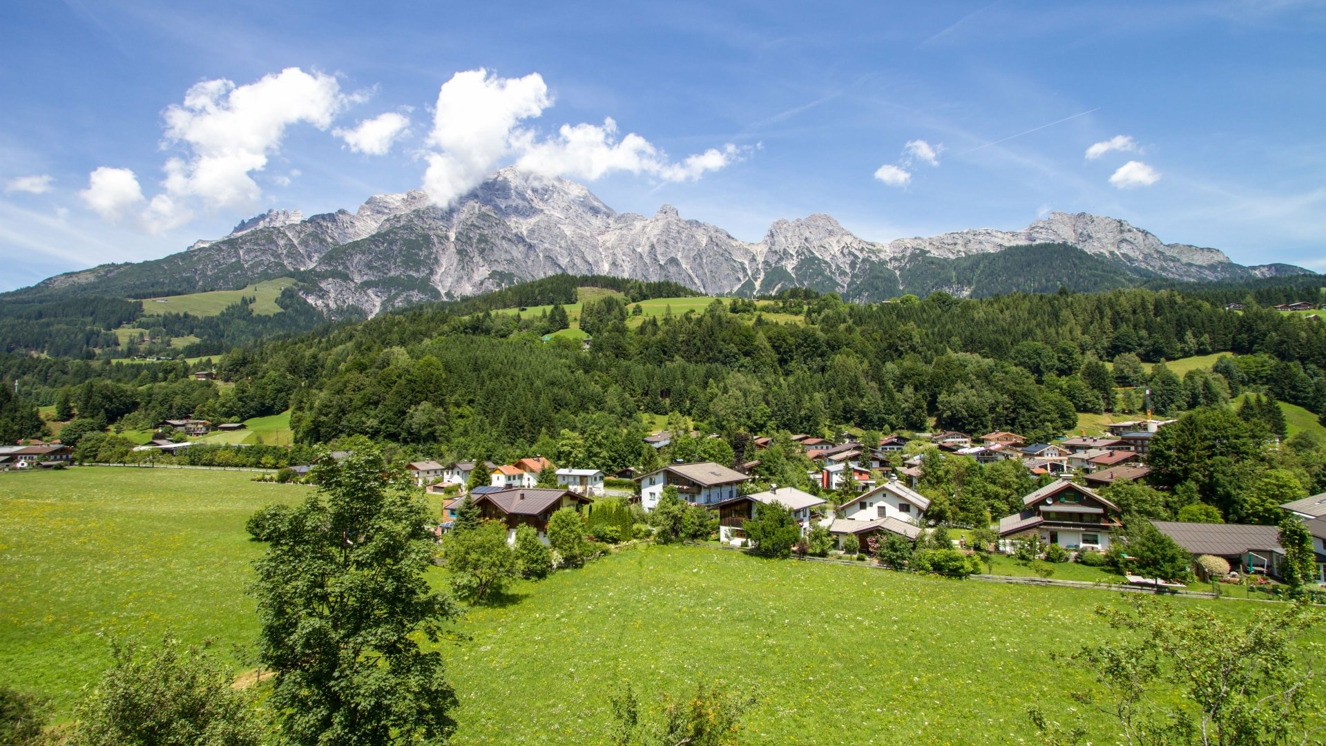 A vibrant 4K Ultra HD man-made village nestled in lush green fields with towering mountains under a bright blue sky, captured as a stunning PC desktop wallpaper.
