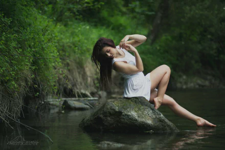 Artistic HD desktop wallpaper featuring a woman in a white dress, gracefully posed on a rock in a tranquil forest stream setting.