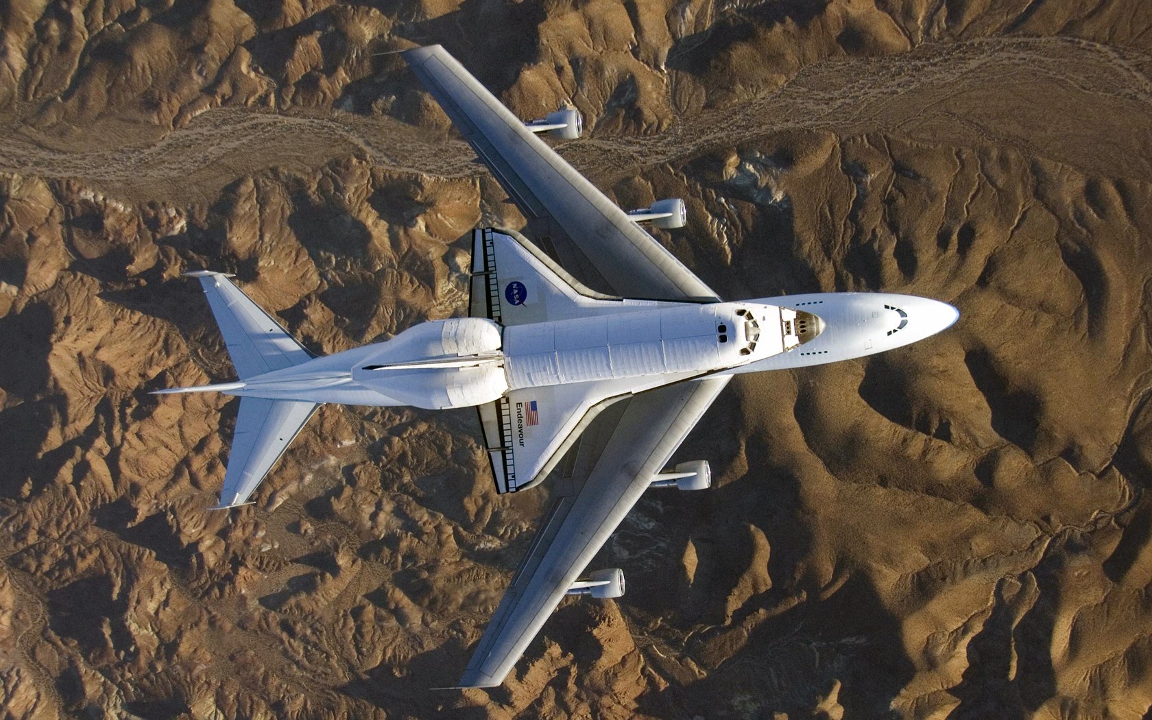 A high-definition desktop wallpaper showing the Space Shuttle Endeavour mounted atop a jumbo jet flying over rugged desert terrain.