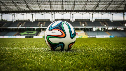 HD desktop wallpaper showing a close-up of a soccer ball on a green field in an empty stadium. The background includes the stadium's seats and partially covered roof.