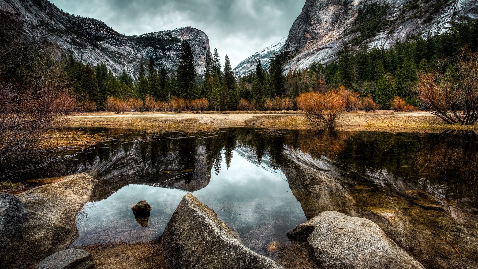 HD PC desktop wallpaper: mountain valley with pine forest and autumn shrubs mirrored in a glassy lake, rocks in foreground — nature reflection.