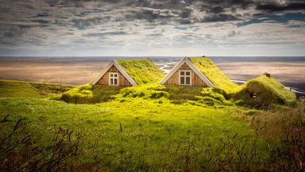 HD PC desktop wallpaper featuring two man-made houses with grass-covered roofs blending seamlessly into a lush green landscape under a dramatic cloudy sky.