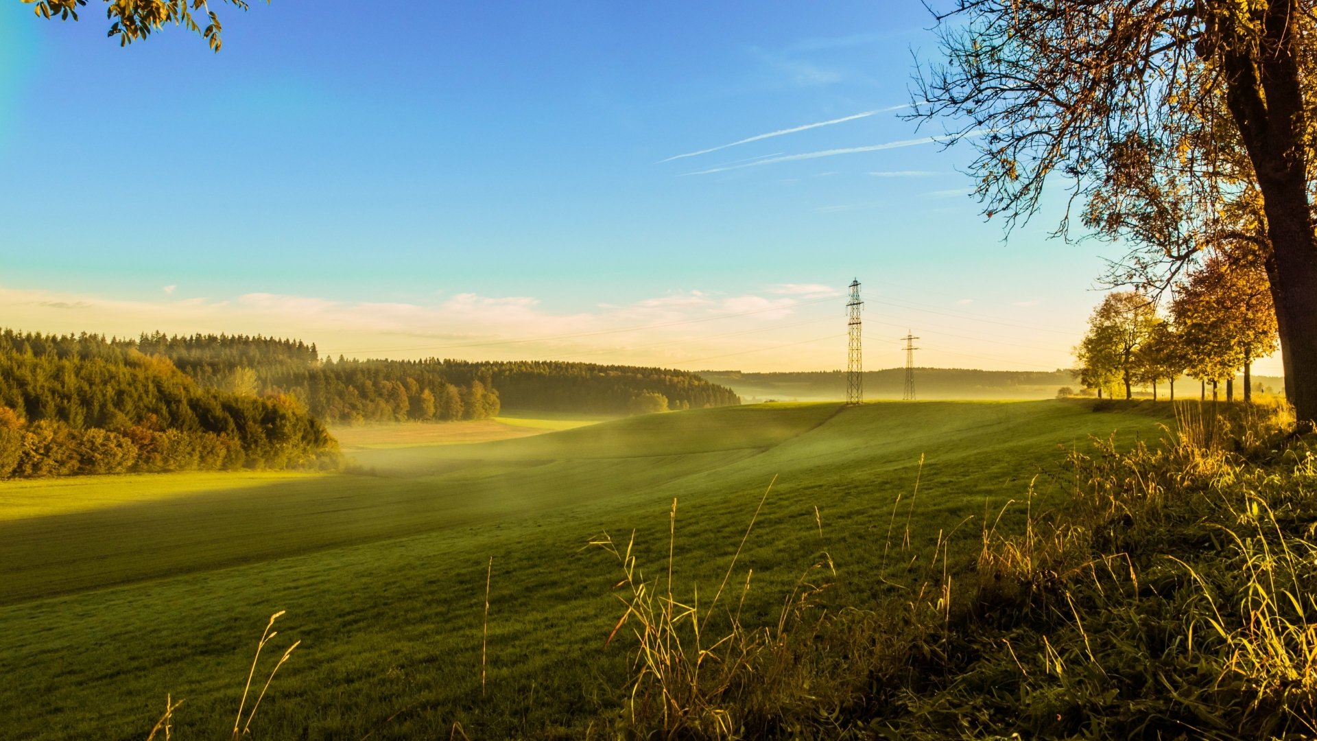 Nature landscape 4K Ultra HD PC desktop wallpaper: sunlit rolling green fields with morning mist, distant forest and power lines under a clear blue sky.