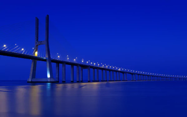 HD desktop wallpaper featuring the illuminated Vasco da Gama Bridge stretching over calm waters against a deep blue evening sky.