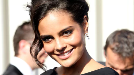 Close-up portrait of a smiling female model in an elegant black dress, soft studio lighting and blurred figures behind, 4K Ultra HD desktop wallpaper.