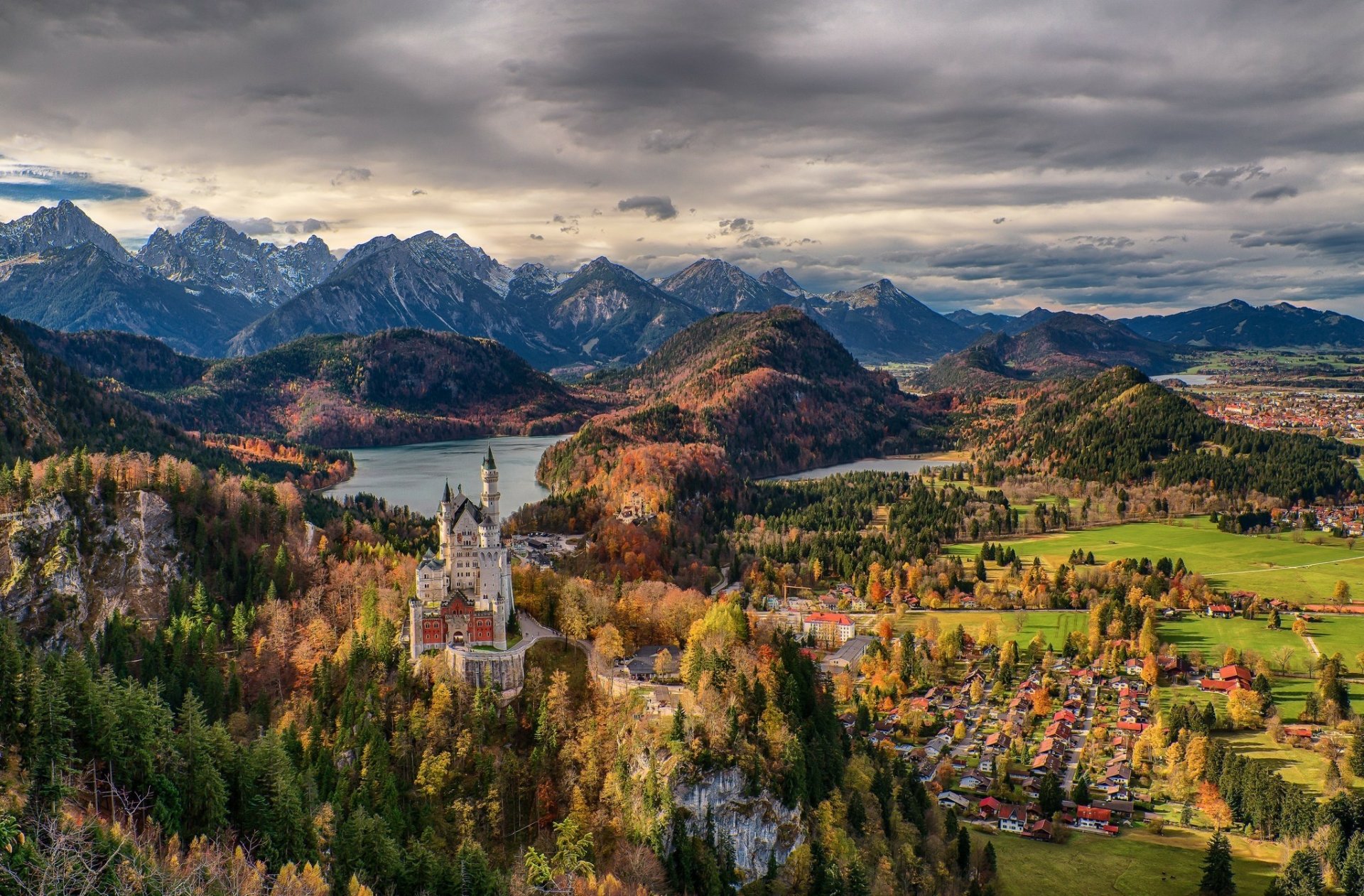 A stunning HD wallpaper of Neuschwanstein Castle in Bavaria, Germany, overlooking a village by a lake with mountains in the background.