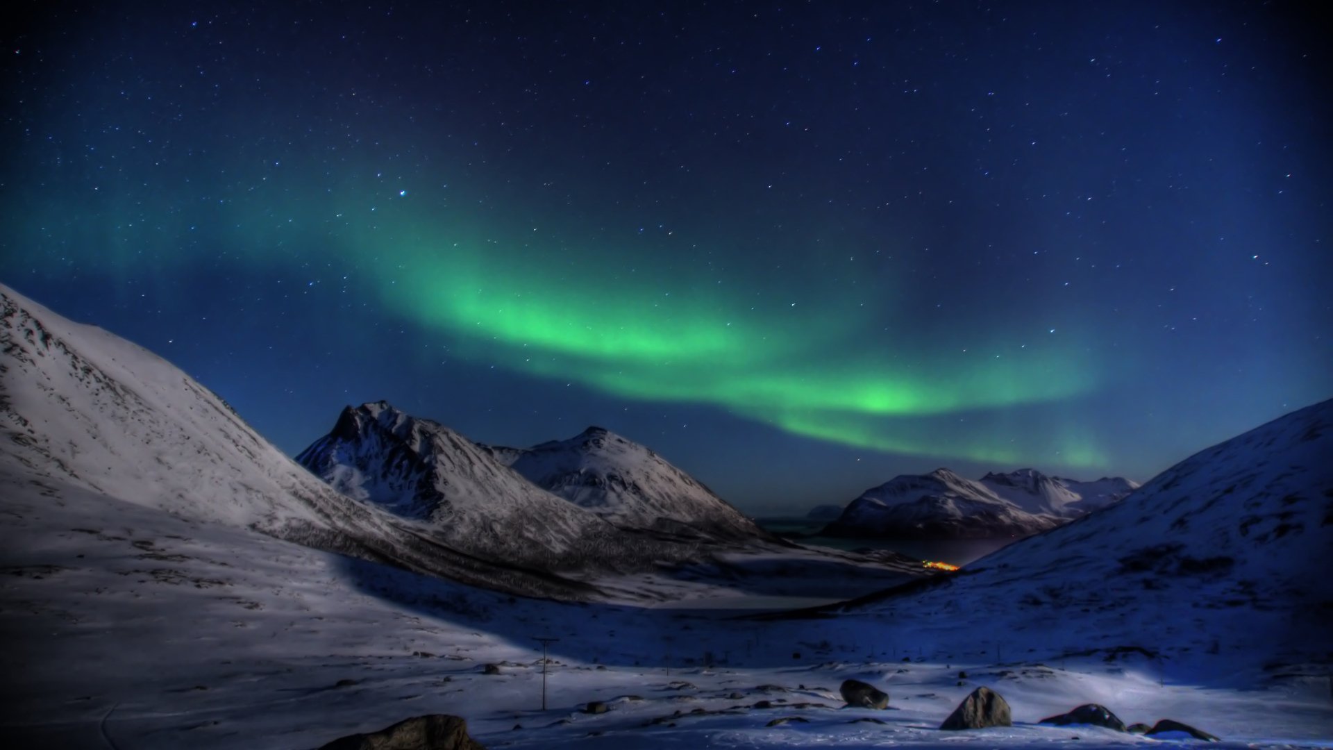 Nighttime snow-covered landscape with the aurora borealis lighting up the sky, set against a backdrop of mountains. This HD desktop wallpaper captures the natural beauty of the northern lights.