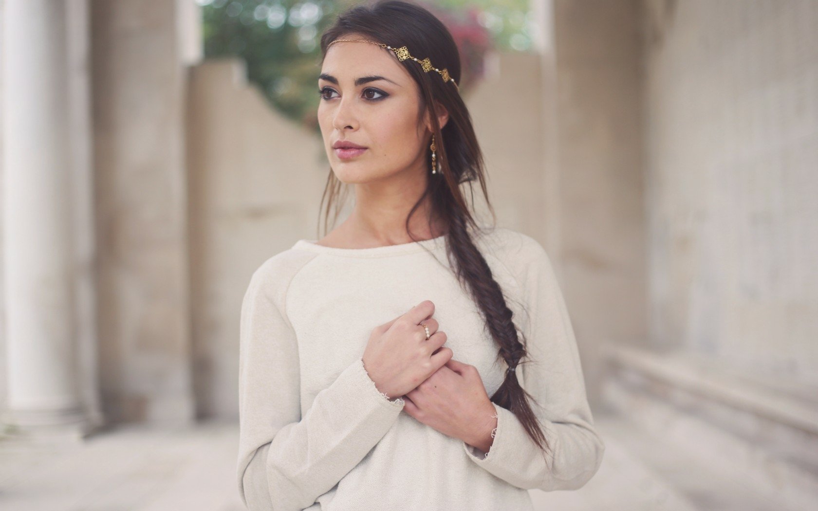 HD desktop wallpaper featuring a brunette woman with a braided hairstyle and floral headband, standing thoughtfully against a blurred architectural background.