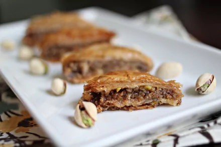 Close-up food shot of pistachio baklava slices on a white plate, syrup-glazed layers and scattered pistachios — 5K Ultra HD PC desktop wallpaper and background.