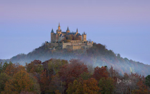 Hohenzollern Castle rises majestically on a mist-covered hill, surrounded by vibrant autumn foliage, creating a stunning backdrop for a desktop wallpaper.