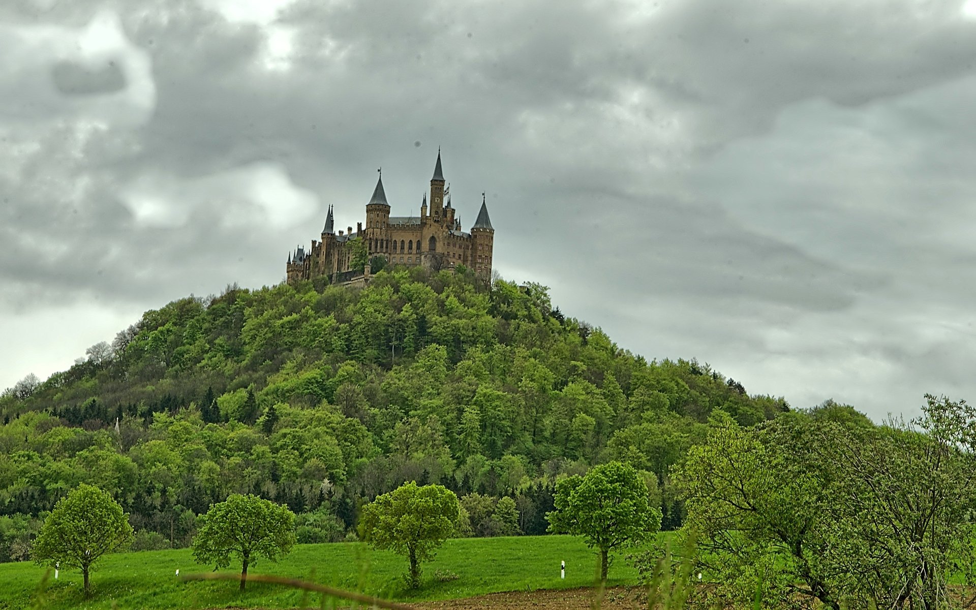 HD desktop wallpaper showcasing the man-made Hohenzollern Castle perched atop a lush green hill under a cloudy sky.