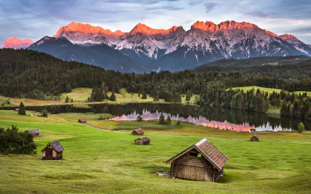 HD landscape photography of a mountain range at sunrise with green fields, scattered wooden cabins, and a reflective lake, designed as a PC desktop wallpaper background.
