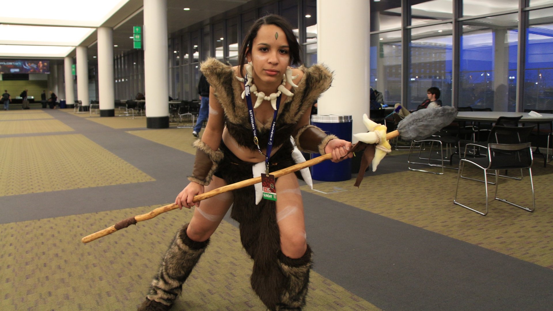 Woman cosplaying Nidalee (League Of Legends) in fur armor and boots, wielding a spear at a convention — 5K Ultra HD PC desktop wallpaper/background.