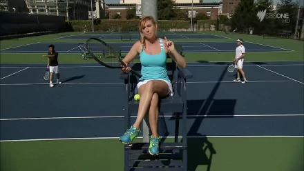 HD desktop wallpaper featuring celebrity Kari Byron seated on a tennis umpire chair, holding a racket on a blue tennis court with players in the background.