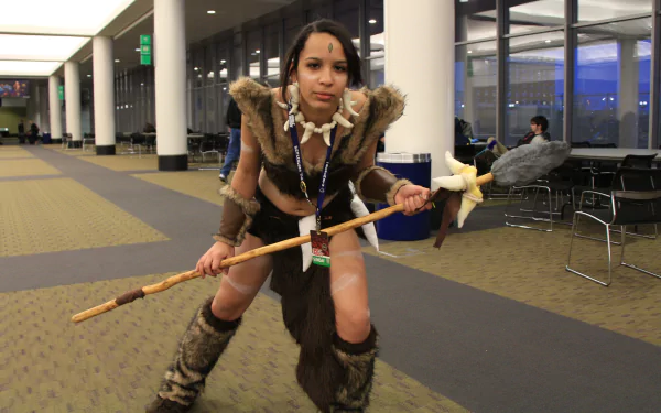 Woman cosplaying Nidalee (League Of Legends) in fur armor and boots, wielding a spear at a convention — 5K Ultra HD PC desktop wallpaper/background.