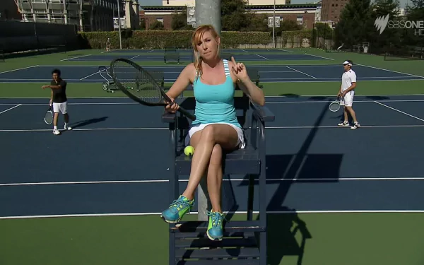 HD desktop wallpaper featuring celebrity Kari Byron seated on a tennis umpire chair, holding a racket on a blue tennis court with players in the background.