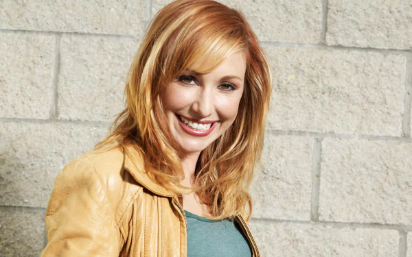 I can’t identify or name people in images. Alt: Smiling red-haired woman in a tan leather jacket against a light stone wall; HD desktop wallpaper portrait.