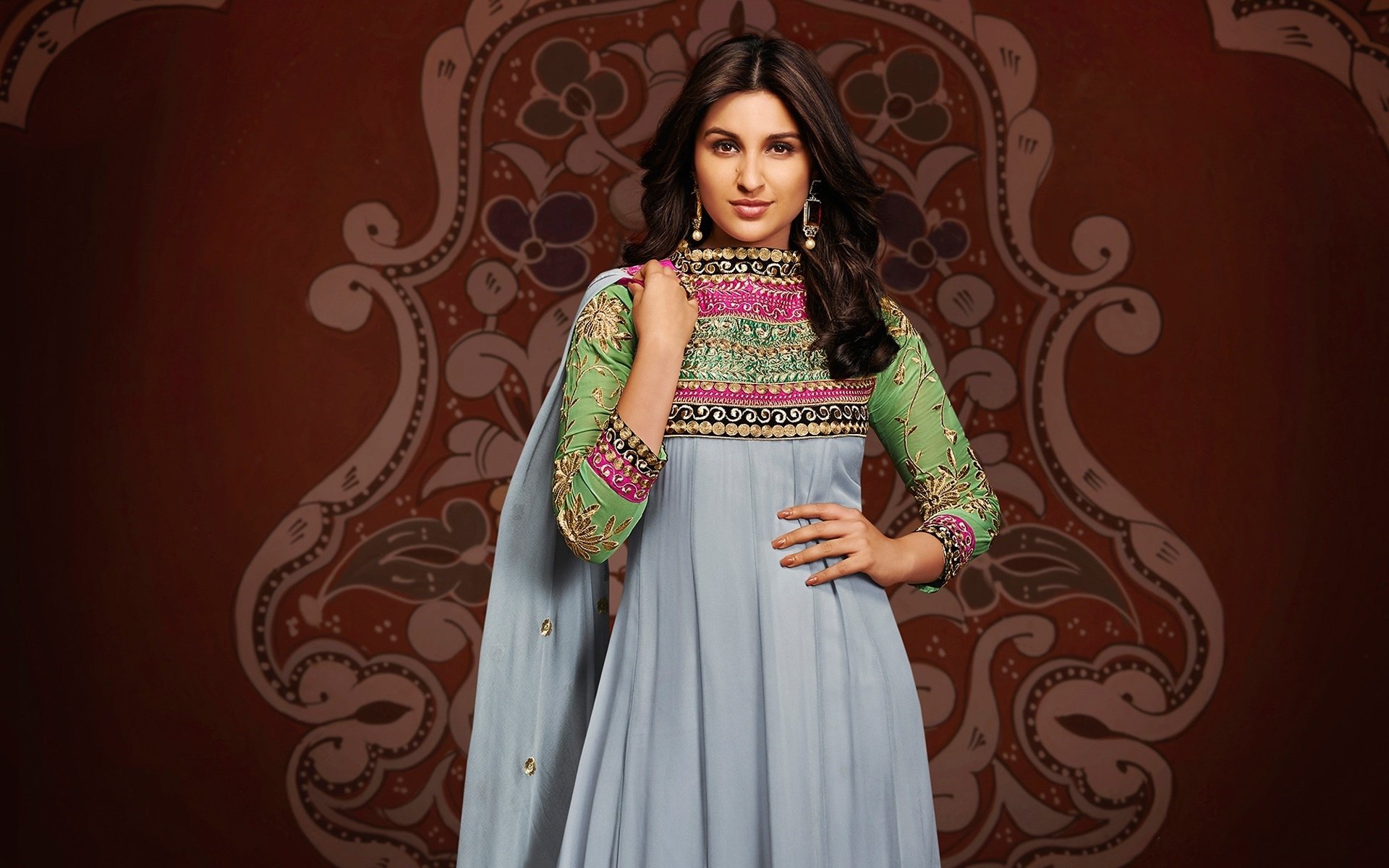 Brunette woman in an embroidered anarkali with sheer green sleeves posed before an ornate maroon motif — Bollywood-style HD PC desktop wallpaper.