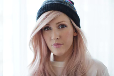 HD wallpaper featuring Ellie Goulding, showcasing her soft pink hair and casual style while wearing a beanie, set against a bright, softly lit background.